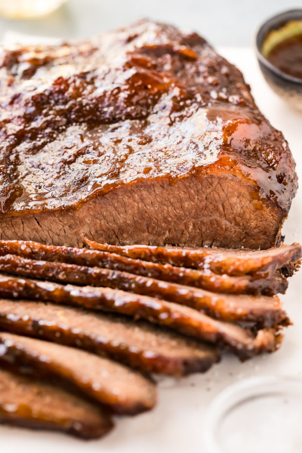 Straight on photo of easy Beef Brisket partially sliced on a white cutting board with a small bowl of salt blurred in the front corner and a small bowl of sauce blurred in the background.