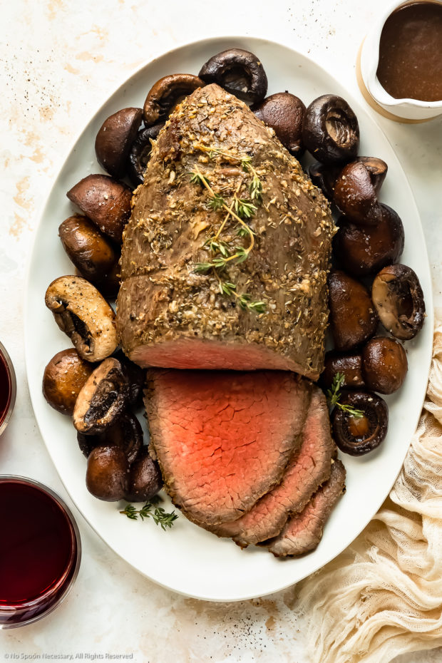 Overhead photo of a eye of round roast beef that's been partially sliced next to a bed of mushrooms on a white platter with a ramekin of gravy and red wine glasses next to the platter.