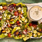 Overhead photo of a garden salad with tomatoes, cucumbers, croutons, and a bowl of buttermilk ranch dressing next to the salad.